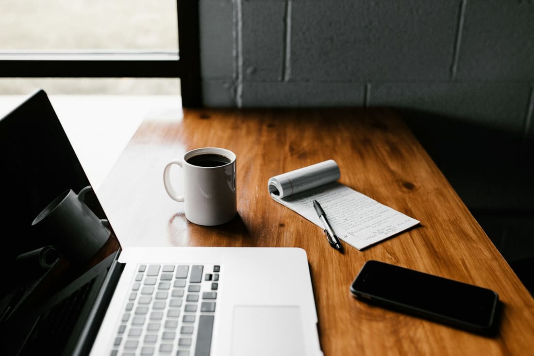 A desk beside a window with a laptop, a mug of coffee and a notebook